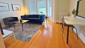 A living room with a dark blue sofa, a small coffee table, a floor lamp, and a ceiling fan. There are two windows with blinds, a radiator, and a potted plant on the windowsill. - Beautifully Set Up 2BR Space (Chicago)