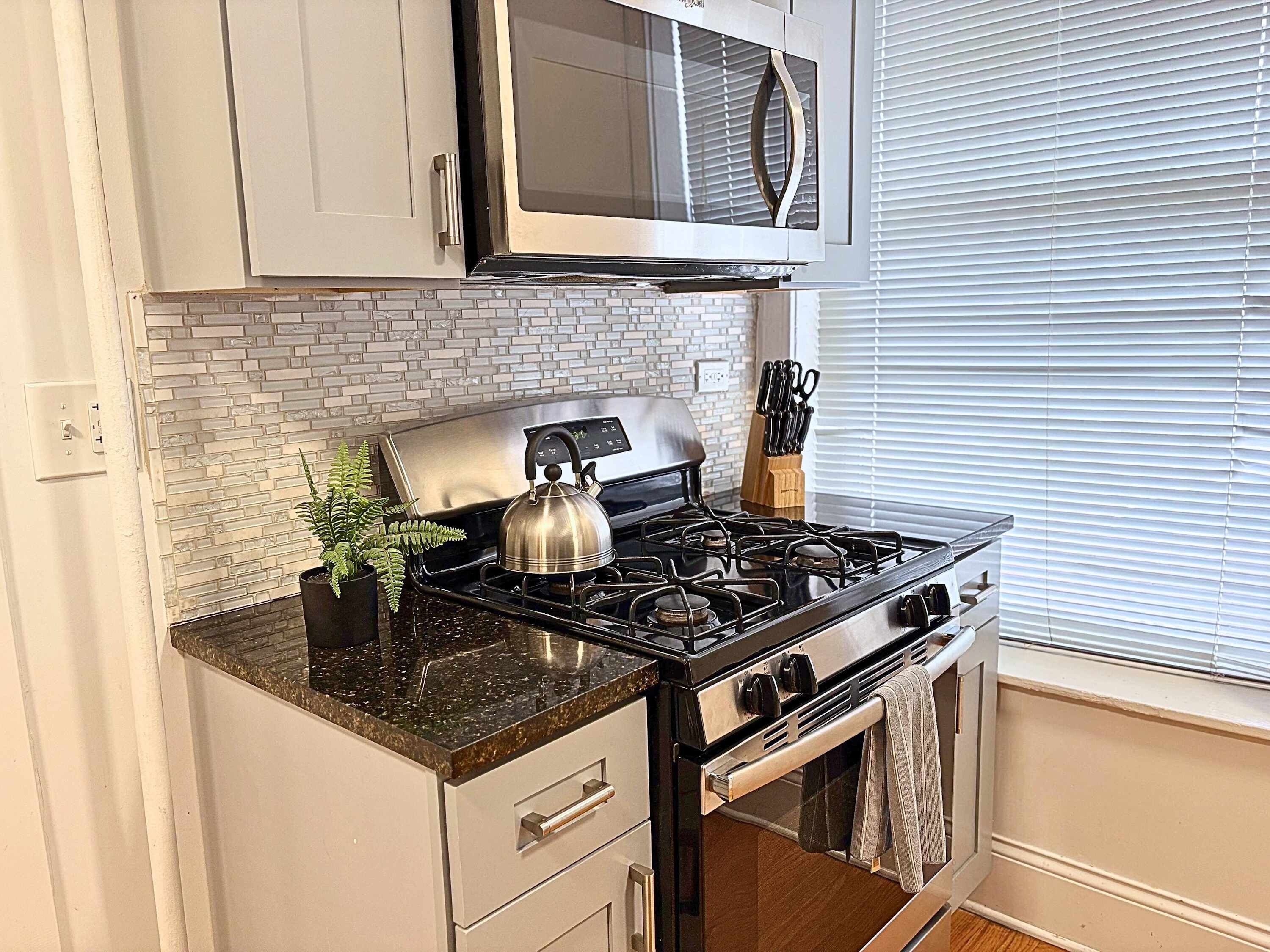 A kitchen with a gas stove, a kettle, a microwave, and a brick backsplash.