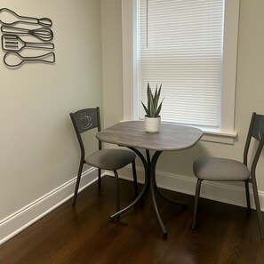 Un área de comedor con una mesa pequeña y dos sillas, una planta en maceta y un utensilio colgante en la pared.