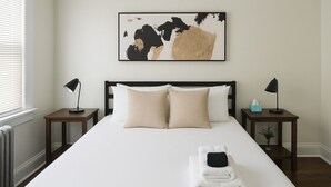 A neatly made bed with two pillows, a bedside table with a lamp, and a framed abstract artwork on the wall. - 1BR Pad with Homey Feel (Chicago)