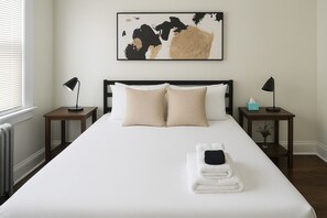 Una cama bien tendida con dos almohadas, una mesita de noche con una lámpara y una obra de arte abstracta enmarcada en la pared.