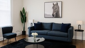 A modern living room with a dark blue sofa, a round coffee table with a vase of white flowers, a potted plant, and a framed picture of a horse on the wall. - 1BR Pad with Homey Feel (Chicago)