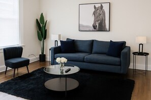 Una sala moderna con un sofá azul oscuro, una mesa redonda de centro con un jarrón de flores blancas, una planta en maceta y un cuadro de un caballo en la pared.