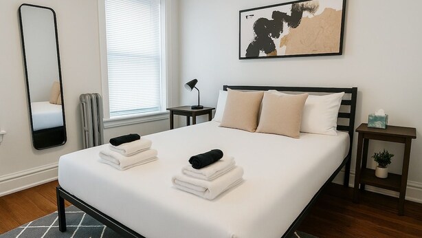 A neatly made bed with folded towels, a bedside table with a lamp, a small plant, and a wall-mounted abstract painting. - 1BR Pad with Homey Feel (Chicago)