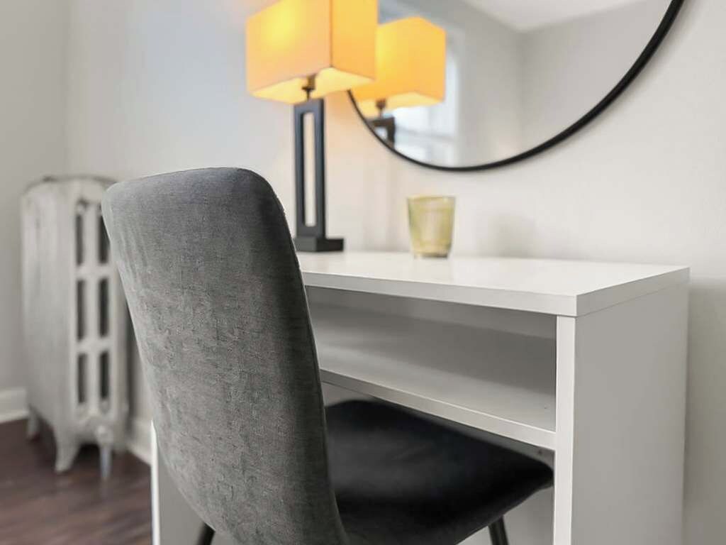 A modern room with a white desk, a grey chair, a lamp, and a mirror.