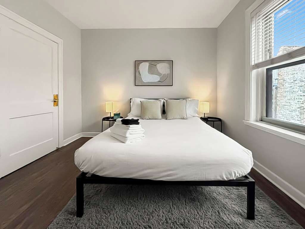 A bedroom with a bed, two bedside tables, a window with blinds, a door, and a wall-mounted abstract painting.