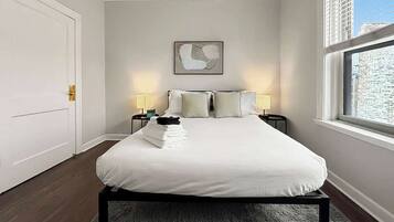 A bedroom with a bed, two bedside tables, a window with blinds, a door, and a wall-mounted abstract painting.