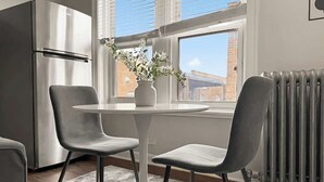 Ett modernt kök med ett kylskåp i rostfritt stål, ett vitt matbord med två grå stolar och ett fönster med utsikt över byggnader.