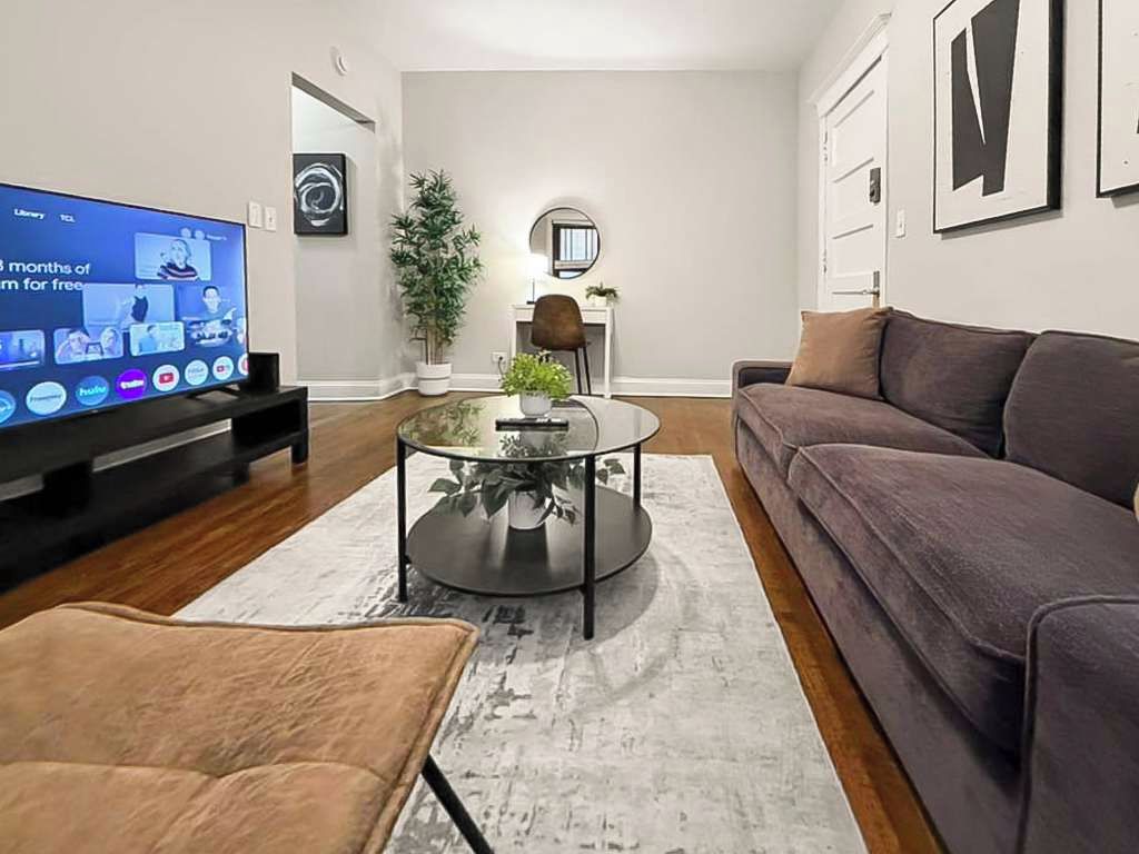 A modern living room with a flat-screen TV, a round glass coffee table, a sofa, and a brown armchair.