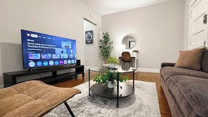Ett modernt vardagsrum med en platt-TV, ett soffbord i glas med växter, en brun läderpall och en grå matta.