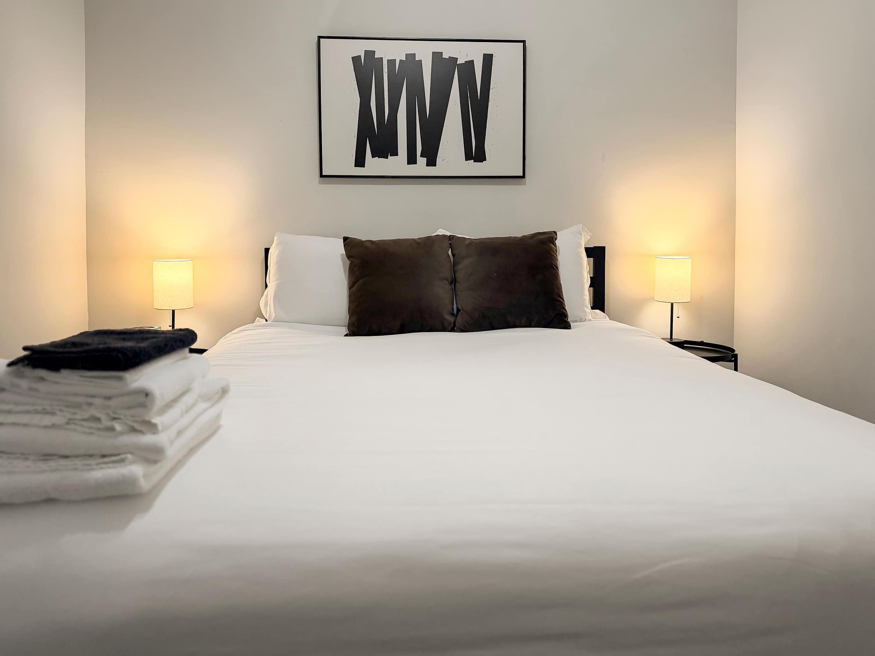 Una cama bien hecha con sábanas blancas y un cojín marrón oscuro, dos lámparas de noche y una obra de arte abstracta enmarcada en la pared.