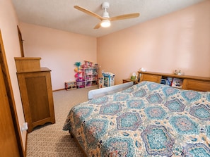 A bedroom with a bed, a wooden wardrobe, a ceiling fan, a toy shelf, and a small desk.