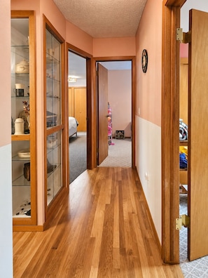A hallway with wooden flooring, glass-fronted cabinets, and a visible bedroom with a bed and a pink toy.