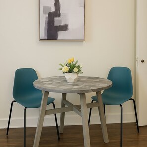 Een ronde tafel met een tafelblad van geometrische tegels, omringd door twee teal stoelen, een witte vaas met gele bloemen en een zwart-wit abstract schilderij aan de muur.