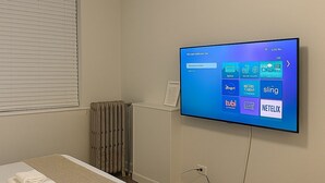 A hotel room with a bed, a wall-mounted television displaying a digital interface, a radiator, and a window with blinds. - Trendy Studio Apartment in Chicago (Chicago)