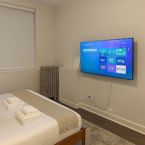 Hotelkamer met een bed, een aan de muur bevestigde televisie met een digitaal scherm, een radiator en een raam met zonwering.