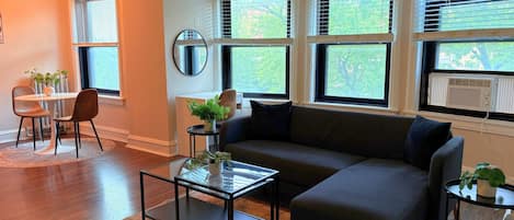Una sala de estar con un sofá modular, una mesa de centro de vidrio, una mesa redonda con sillas y ventanas amplias con persianas.