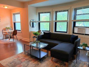 A living room with a sectional sofa, a glass coffee table, a round table with chairs, and large windows with blinds. - Bright 1BR Unit in a Prime Spot (Chicago)