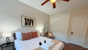 Un lit bien fait, avec du linge de lit blanc et un plaid beige, un ventilateur de plafond en bois, un miroir fixé au mur et une porte, dans une chambre d’hôtel.