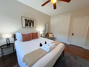 Een opgemaakt bed met witte lakens en een beige plaid, een houten plafondventilator, een spiegel aan de muur en een deur in een hotelkamer.