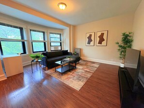 Een moderne woonkamer met houten vloeren, een zwarte bank, een glazen salontafel en een groot raam met zonwering.