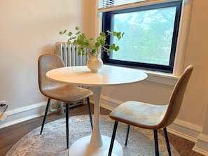 Een kleine ronde tafel met twee stoelen, een bloempot en uitzicht op bomen door een raam.