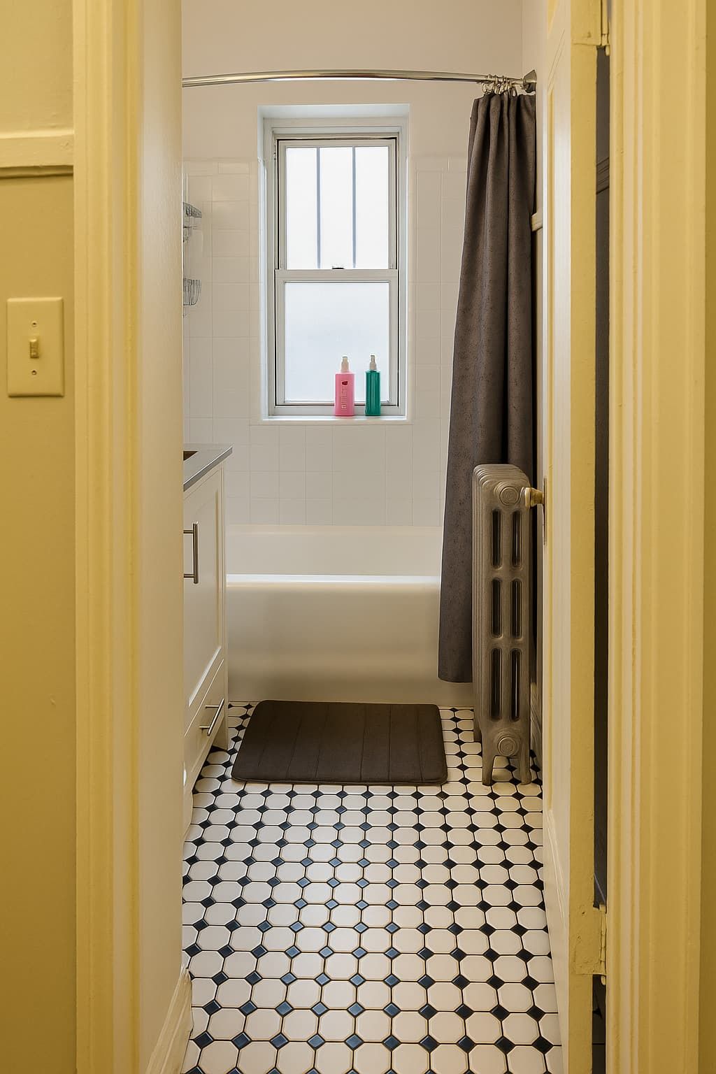 A bathroom with a bathtub, a window with two bottles on the sill, a radiator, and a patterned floor.