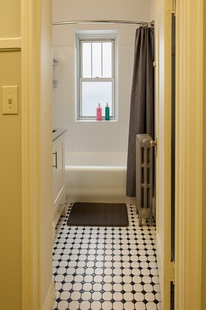 Une salle de bain avec une baignoire, une fenêtre sur le rebord de laquelle sont posées deux bouteilles, un radiateur et un sol à motifs.