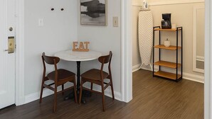 En matplats med ett runt bord och två stolar, en kaffemaskin på en hylla och en inramad tavla på väggen.
