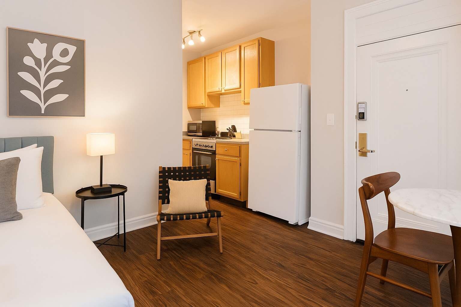 Habitación de hotel compacta con cama, una silla, una mesita, escritorio, kitchenette con armarios de madera, refrigerador blanco y puerta con cerradura electrónica.