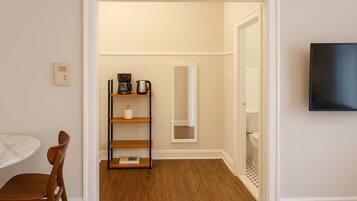 Una habitación con piso de madera, pared blanca, un estante de madera con cafetera, una silla blanca y un televisor montado en la pared.