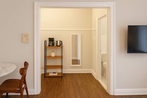 A room with a wooden floor, a white wall, a small wooden shelf with a coffee maker, a white chair, and a TV mounted on the wall. - Cozy Charm Studio Apt in Chicago (Chicago)