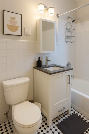Et badeværelse med toilet, vask og badekar. Et billede hænger på væggen, og der er et brusehoved over badekarret.