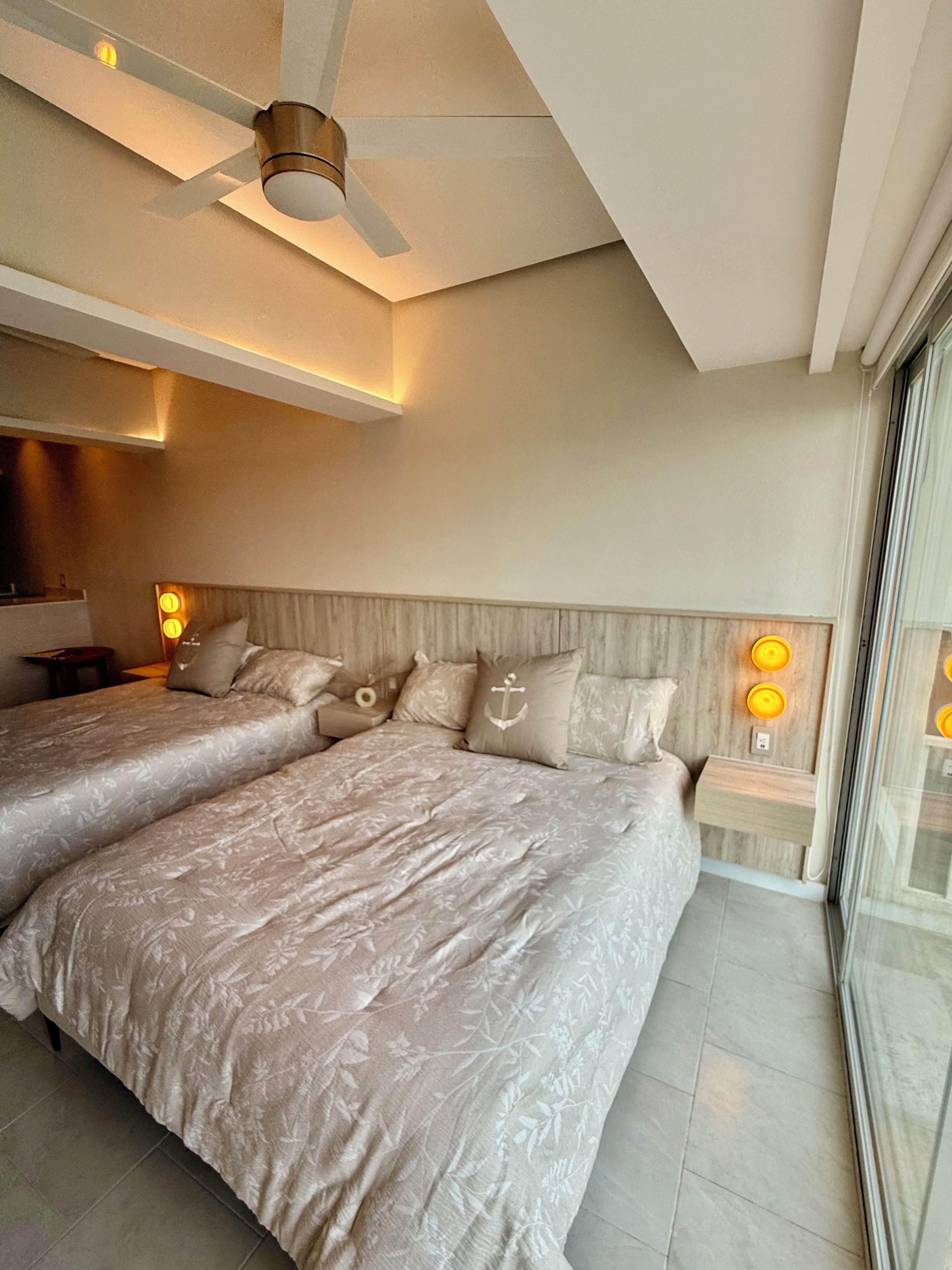 Quarto de hotel com duas camas, ventoinha no teto, abajures e uma janela grande.