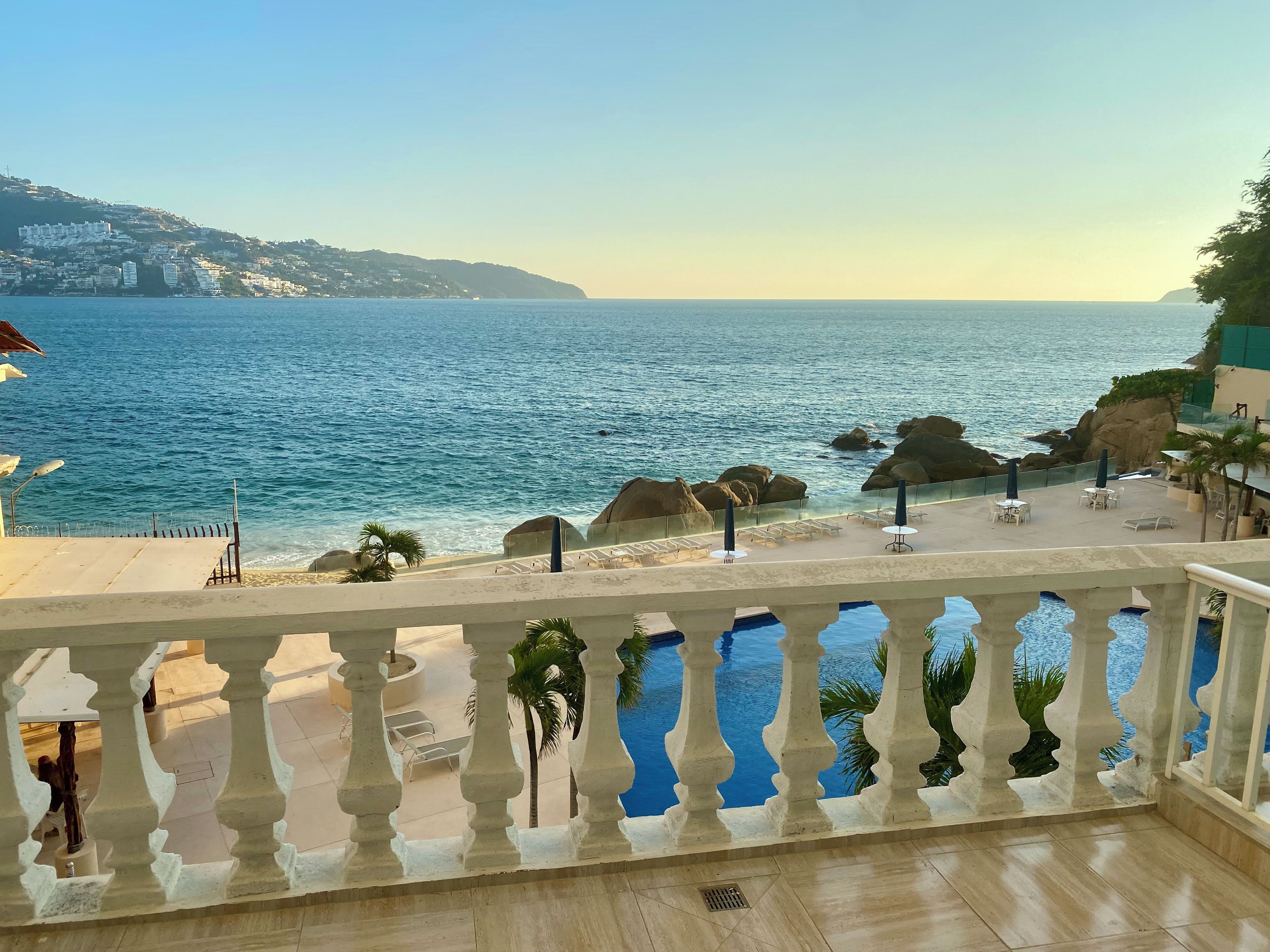 A balcony with a view of the sea, a pool, and a distant mountain range.