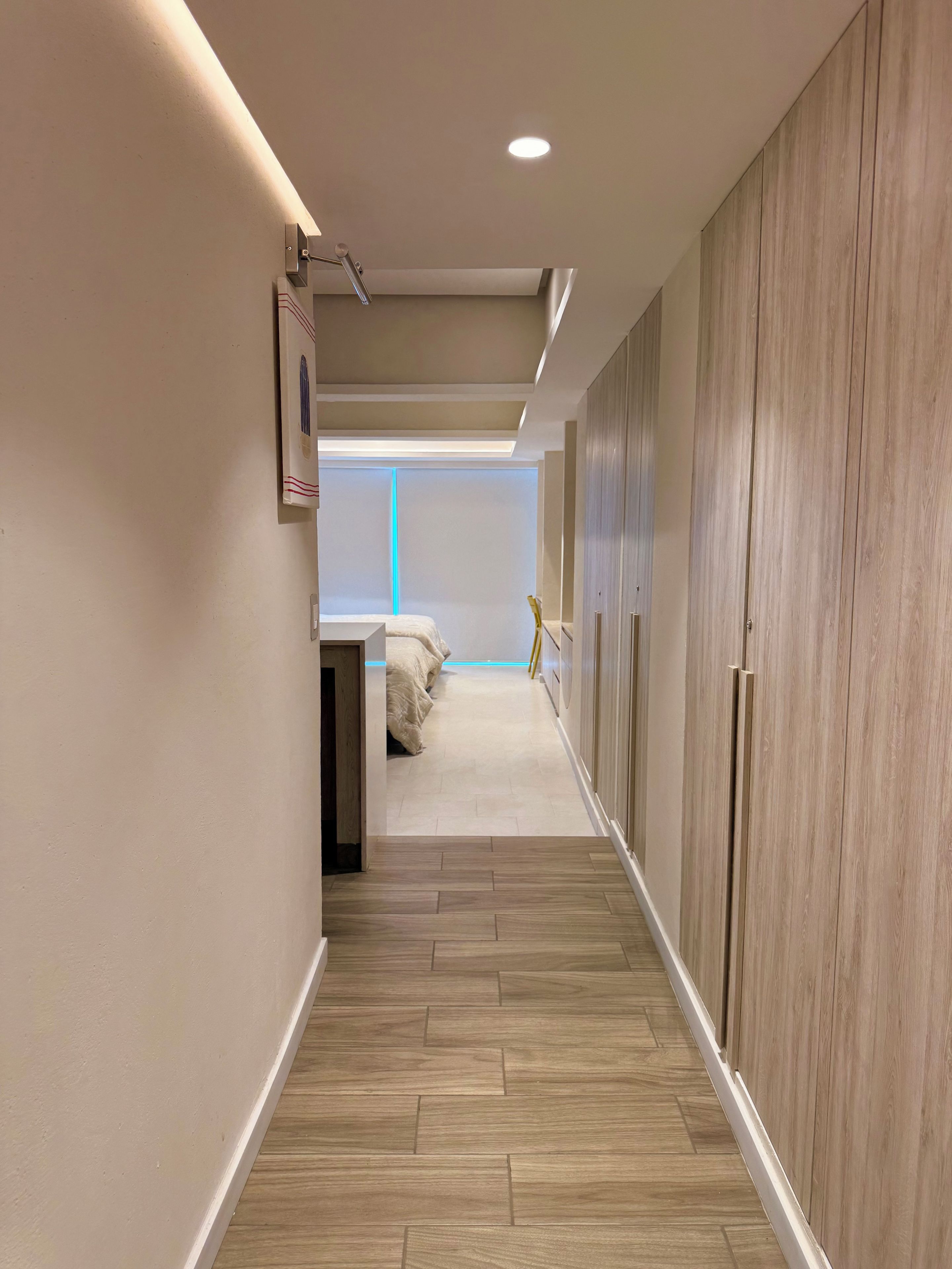 A hallway with a wooden floor, a bed with a white bedspread, and a wardrobe with a light wood finish.