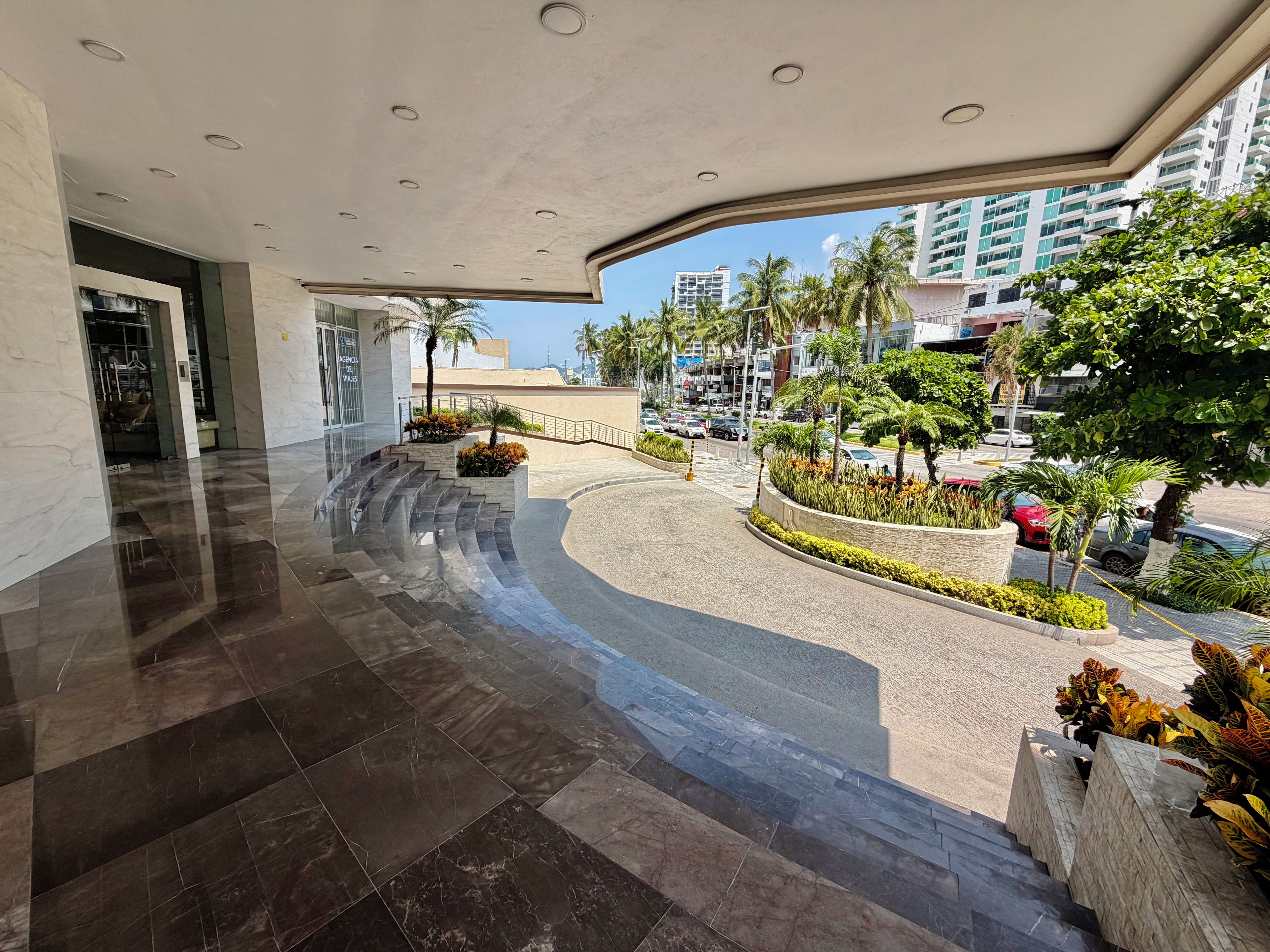 A modern hotel lobby with a curved floor design, a staircase leading to an upper level, and a view of the street with palm trees and parked cars.