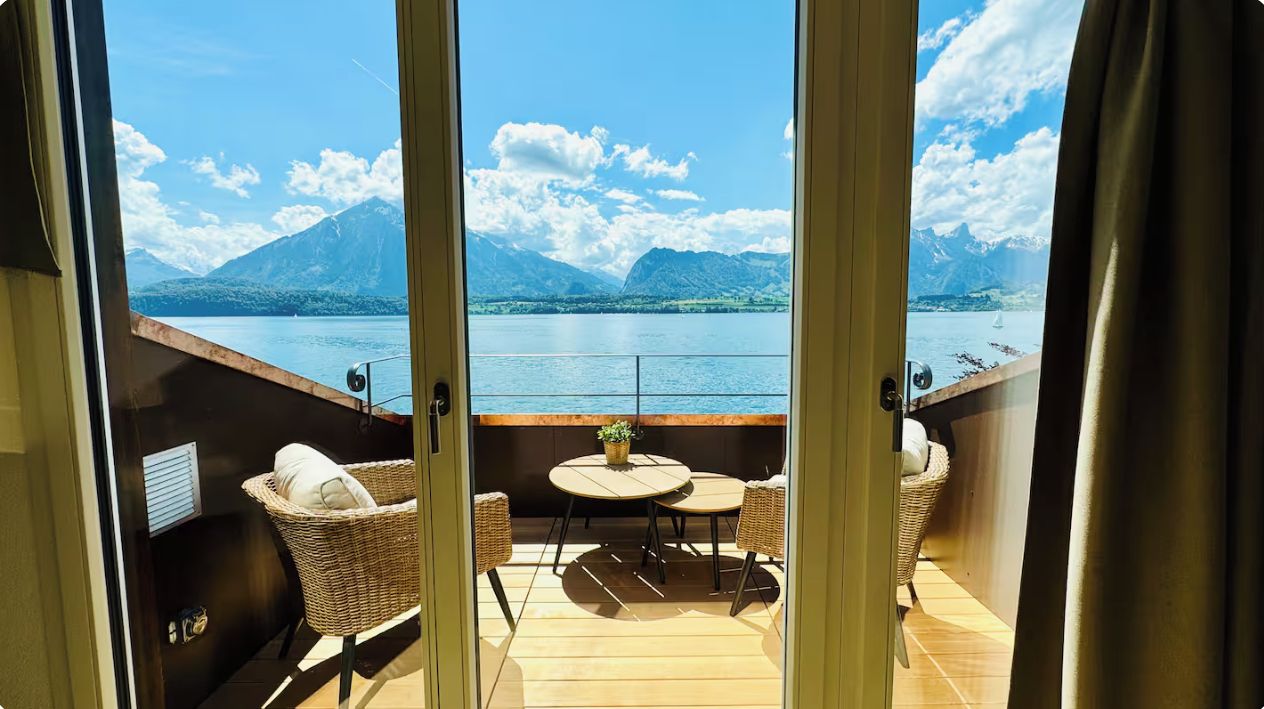 A balcony with wicker chairs and a table, overlooking a lake with mountains in the background.