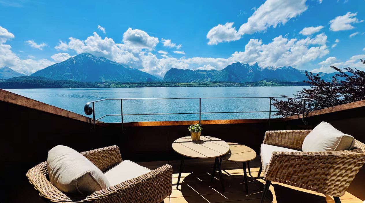 A terrace with wicker furniture and a table, overlooking a lake with mountains in the background.