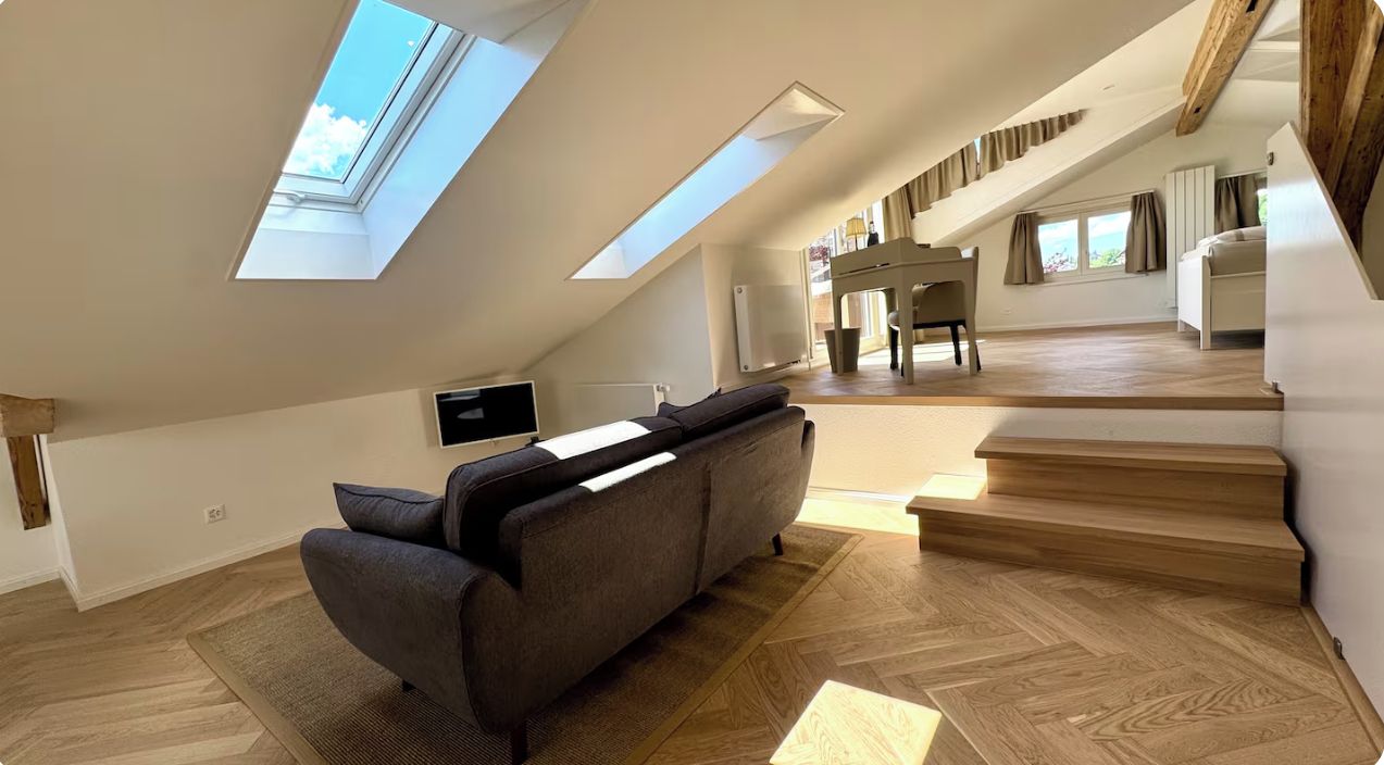 A spacious loft-style room with a high ceiling, skylights, a sofa, a piano, and a staircase.