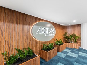 A reception area with a wooden wall, a circular sign with 'AEGEAN Apartments' text, and potted plants. - High-Rise 2-Bed Apartment in Surfers Paradise (Surfers Paradise)