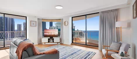 A modern living room with a large flat-screen TV, a sofa, a coffee table, and a view of the city and ocean through floor-to-ceiling windows.