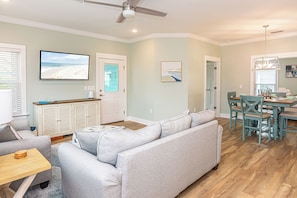 Ein Wohnzimmer mit grauem Sofa, einem Holzesstisch mit Stühlen, einem Deckenventilator und einem Fernseher an der Wand.