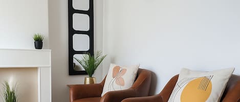 Ett vardagsrum med två bruna läderfåtöljer, en vit öppen spis, en klocka och en geometrisk väggdekoration.