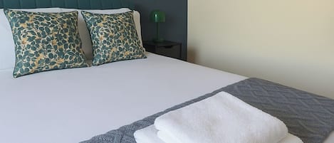 A neatly made bed with a grey knitted blanket and two white towels, a green lamp on a bedside table, and patterned pillows.
