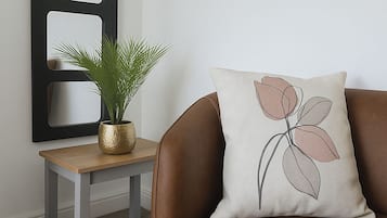 Un sofá de cuero marrón con un cojín decorativo de diseño floral, una mesita de madera pequeña con una planta en maceta, y una obra de arte mural negra con secciones recortadas.