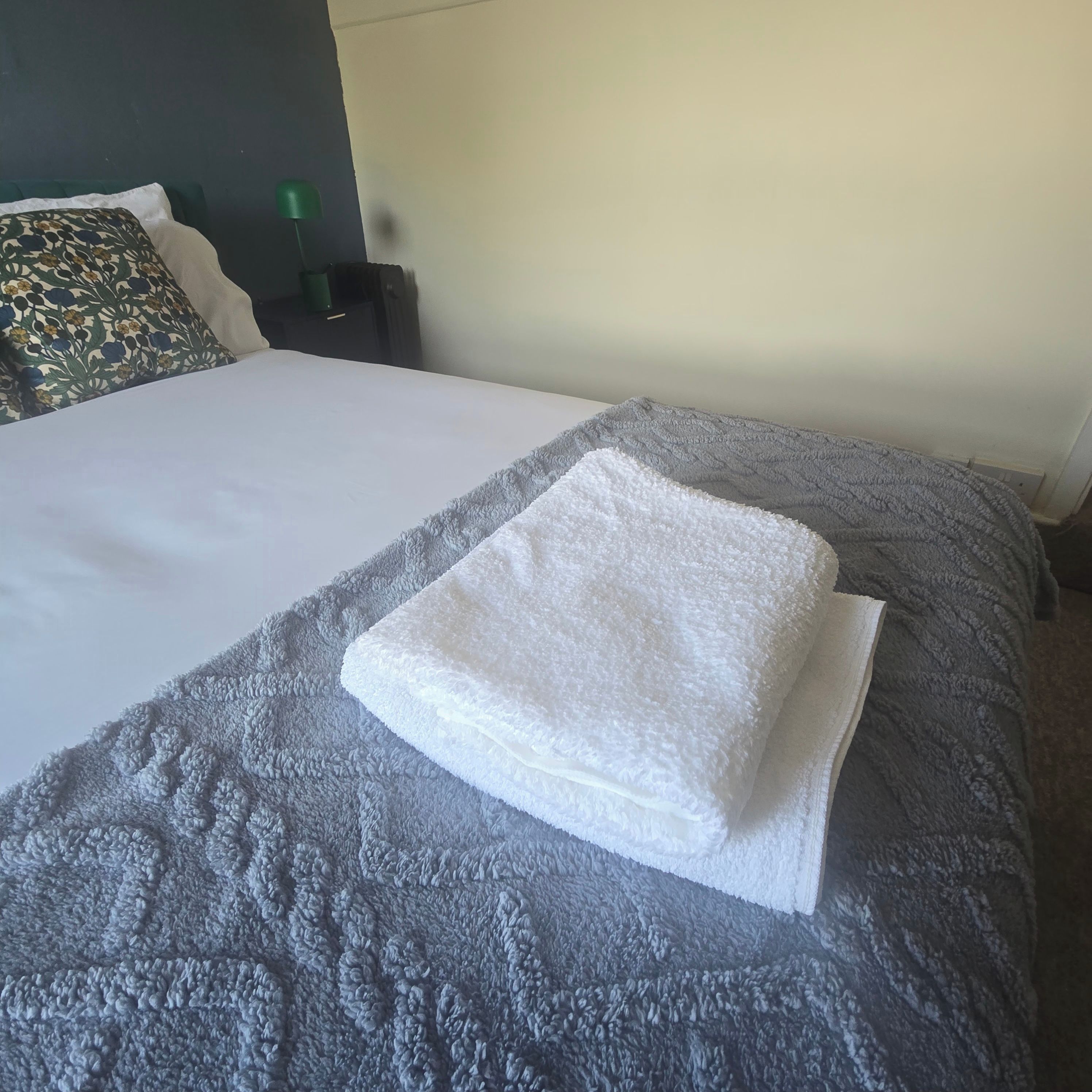 Un lit bien fait, avec une serviette blanche pliée posée sur le dessus, une couverture grise à texture et un coussin décoratif orné d’un motif floral.