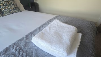 Un lit bien fait, avec une serviette blanche pliée posée sur le dessus, une couverture grise à texture et un coussin décoratif orné d’un motif floral.