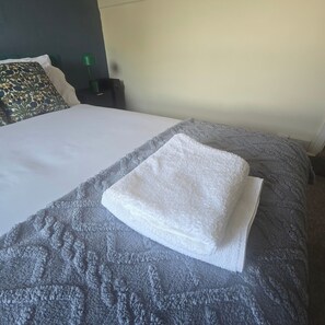 A neatly made bed with a folded white towel on top, a grey textured blanket, and a decorative pillow with a floral pattern.