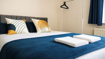 Een net opgemaakt bed met een blauwe deken en twee witte handdoeken, een grijze hoofdeinde en een zwarte kapstok aan de muur.
